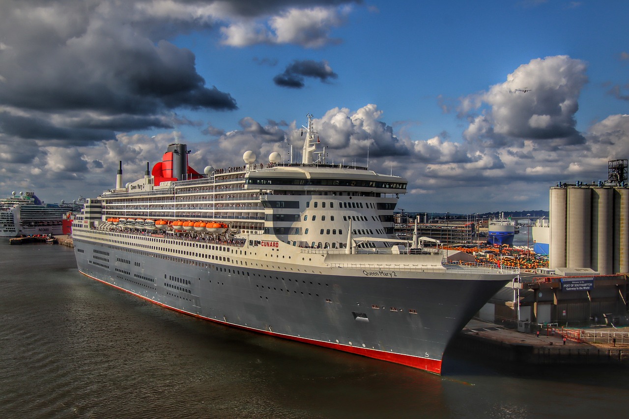 A large cruise ship, Queen Mary 2, docked at Southampton port under a partly cloudy sky. Another cruise ship is visible in the background.